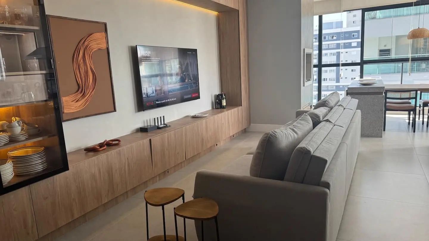 Sala de estar com sofá grande e painel de madeira com TV, cozinha ao fundo