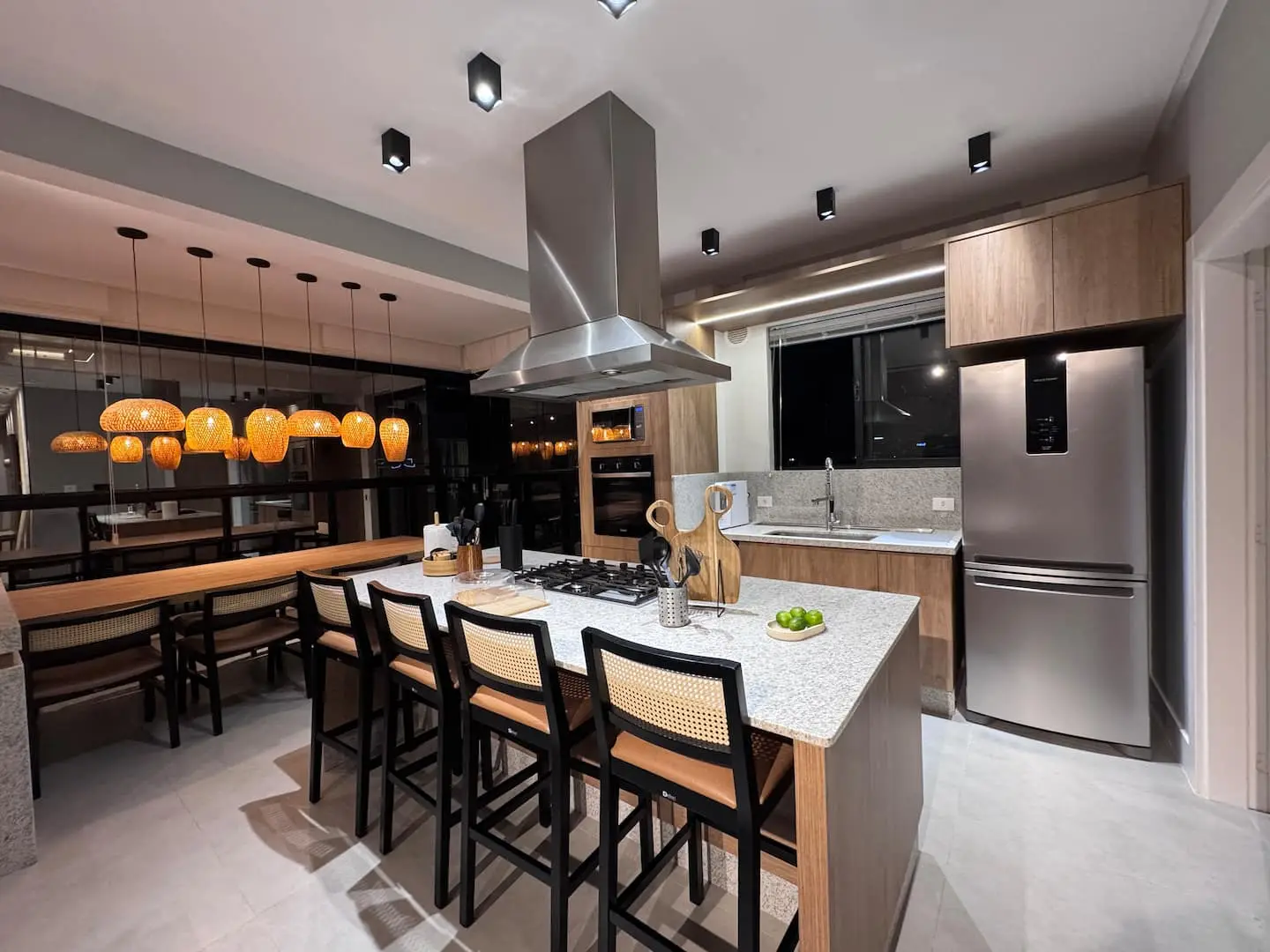 Cozinha gourmet do apartamento à noite, com ilha de pedra, mesa de madeira e pendentes de palha acesos
